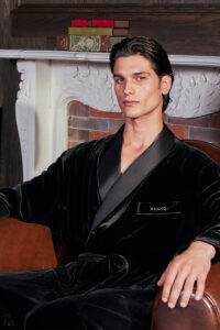 Model seated in an armchair wearing the MANITO velvet robe for men, showcasing its rich black sheen and shawl collar.