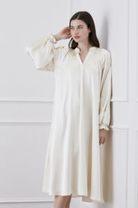 Woman in creamy white midi dress against white background, elegantly touching hair to showcase lantern sleeves.