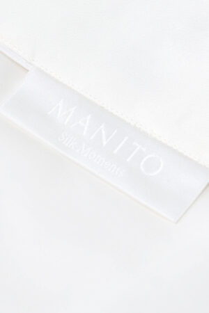 Corner of Gleam Silk Quilt showing the MANITO Silk Moments label and fine stitching on crisp white silk.